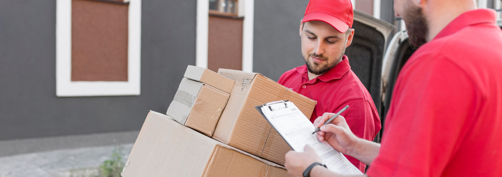 Two delivery workers in red uniforms unload cardboard boxes from a van; one writes on a clipboard nearby.